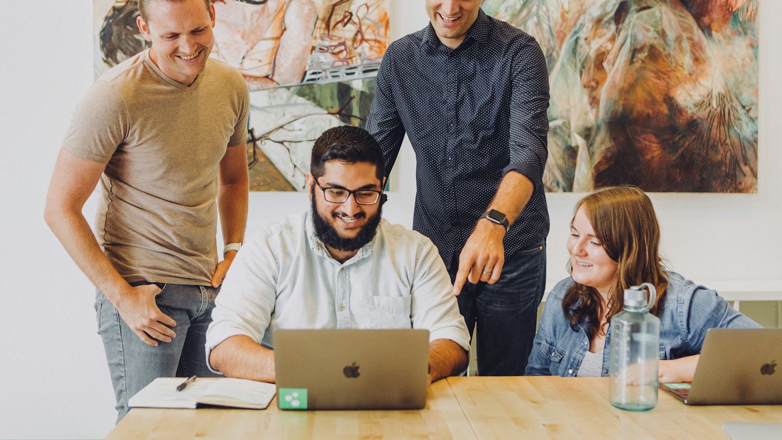 Four coworkers smiling together while looking at a laptop in a bright office